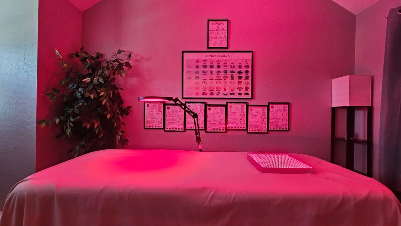 The session room in soft pink light, with a padded table and a wall of framed chakra charts